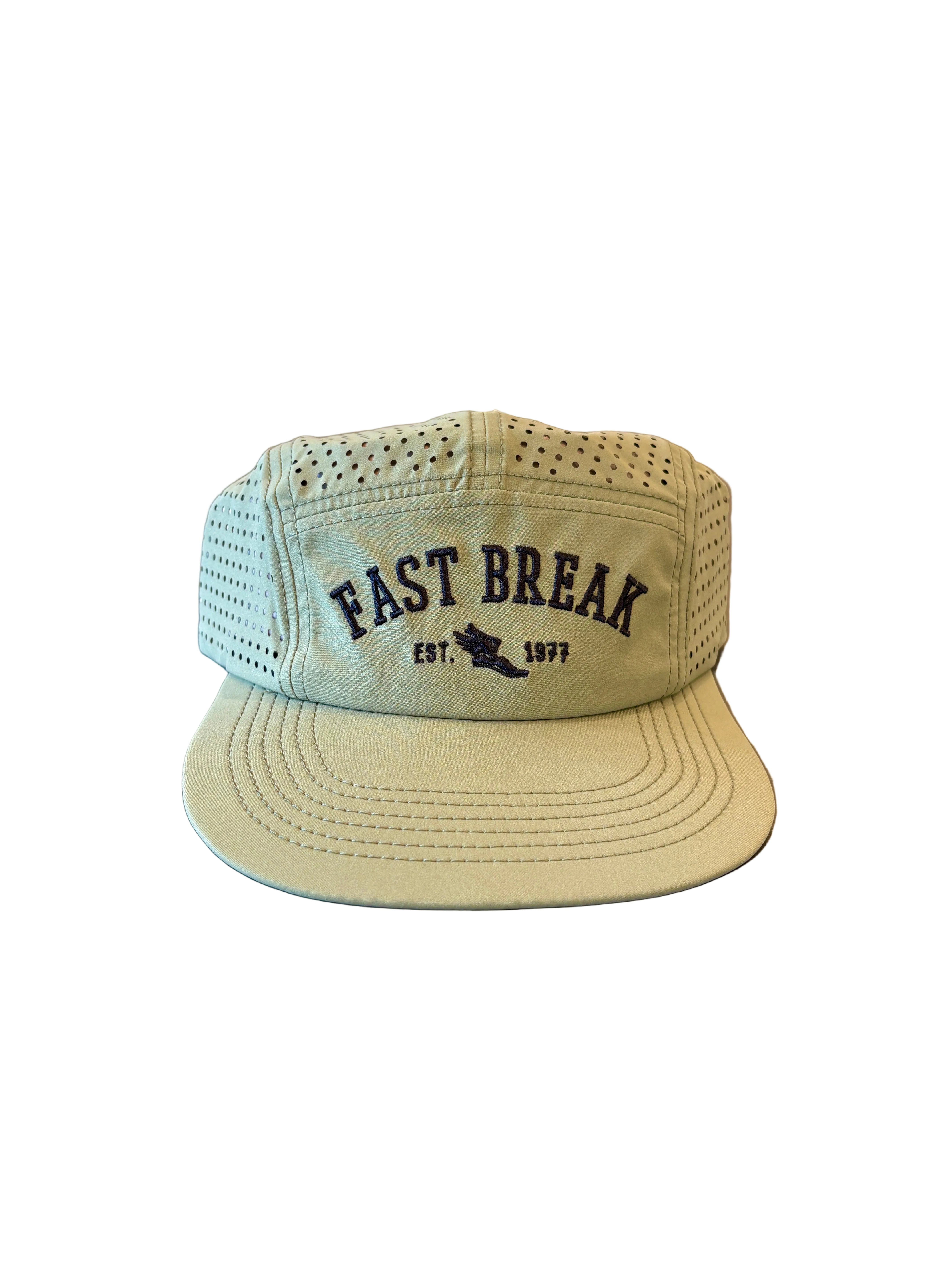 A light sage green mesh trucker cap with 'FAST BREAK EST. 1877' printed in navy on the front, resting on a dark gray table with a blurred shoe display in the background.